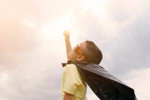 boy in a cape and sunglasses raising his fist to the clouds as he pretends to be a superhero