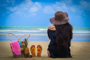 woman sitting on beach wearing a black cover-up and gray floppy hat next to a pineapple, flip flops, and bag as she looks out at the ocean